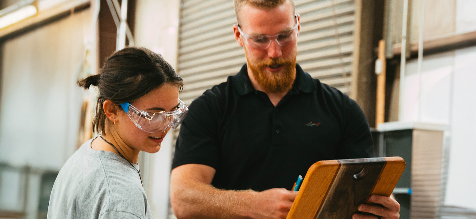 A student talking to a coach while working on a project. The coach is holding a clip board